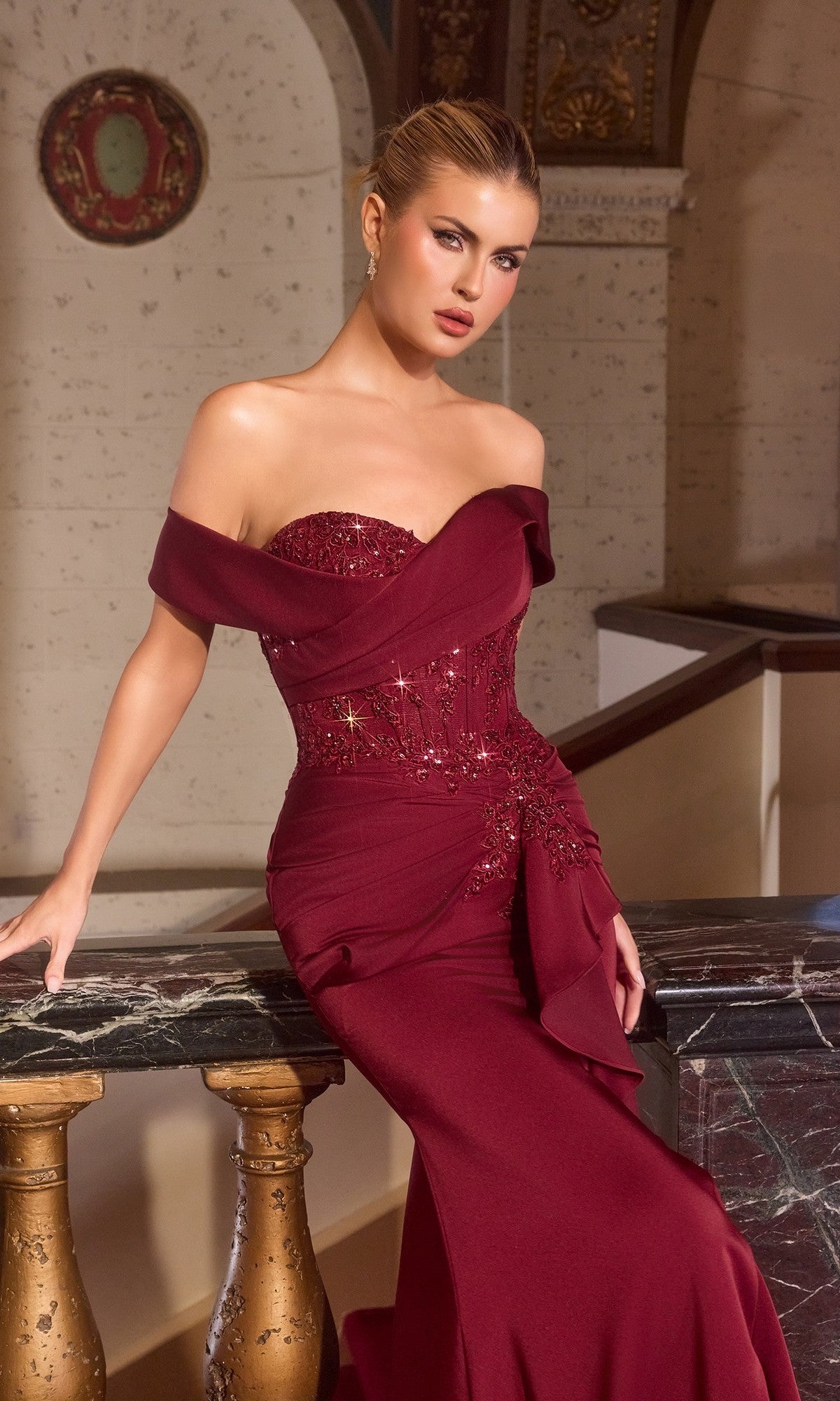 Woman wearing the Ladivine CM387 long formal dress, a fitted off-the-shoulder gown with a beaded bodice, standing by a marble and brass railing in an ornate interior.